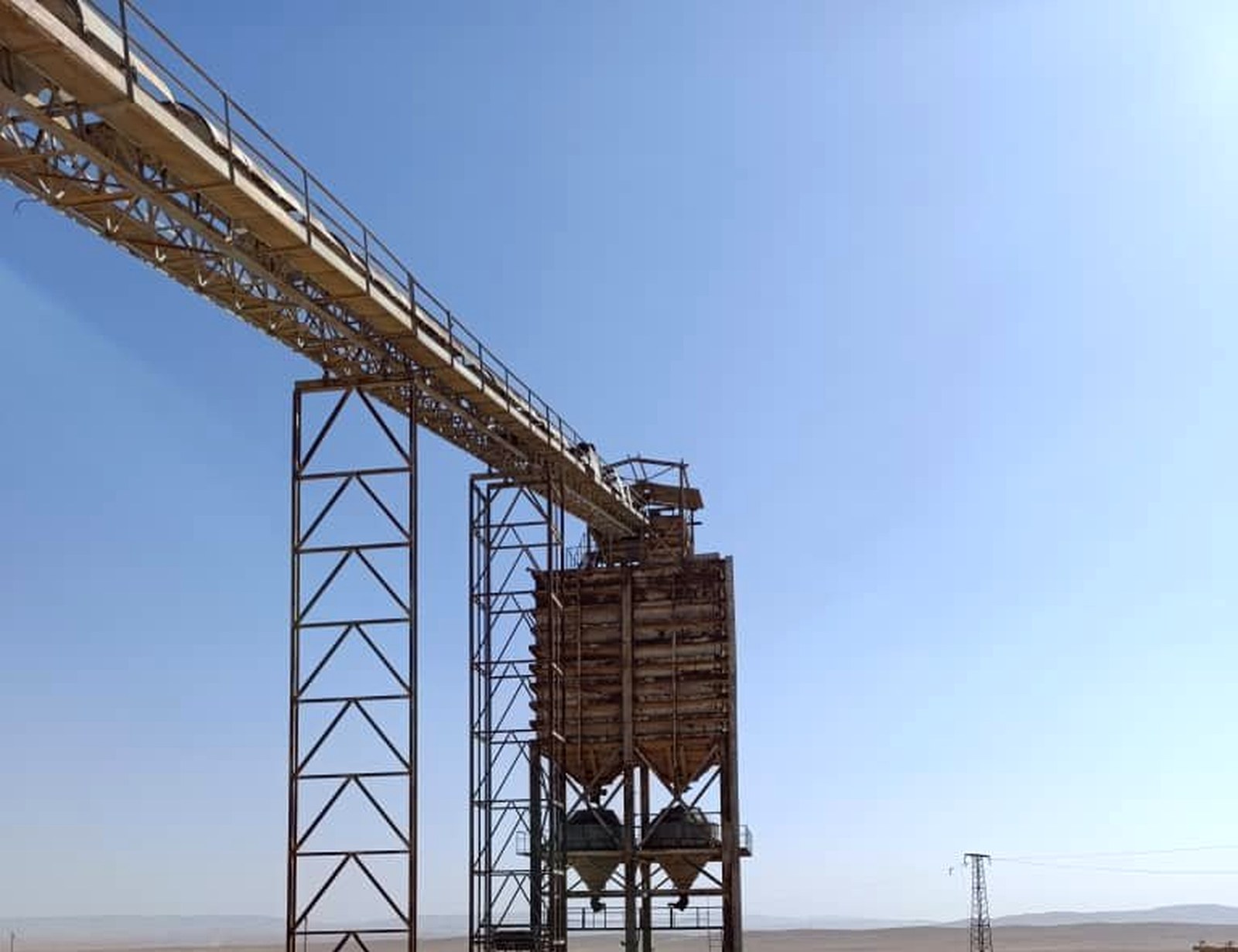 Existing industrial infrastructure at the project site: conveyor system, gravity-feed hoppers, and structural framework against desert horizon